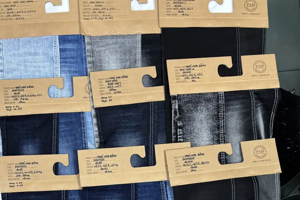Nhiều mẫu vải denim xanh, xám và đen khác tông xếp cạnh nhau tại kho Phúc Anh Đăng, mỗi mẫu kẹp một tag giấy kraft ghi mã hàng, thành phần và khổ vải