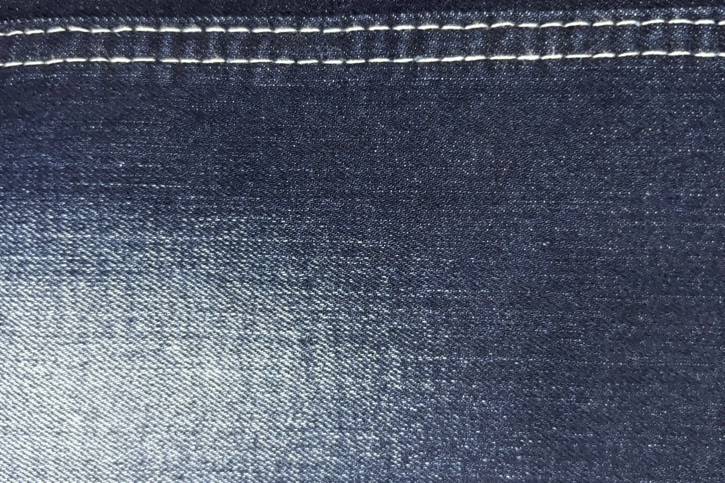Cận cảnh mặt vải denim xanh chàm với đường chỉ may trắng chạy ngang phía trên và một dải vệt bạc sáng nổi rõ trên nền vải đậm