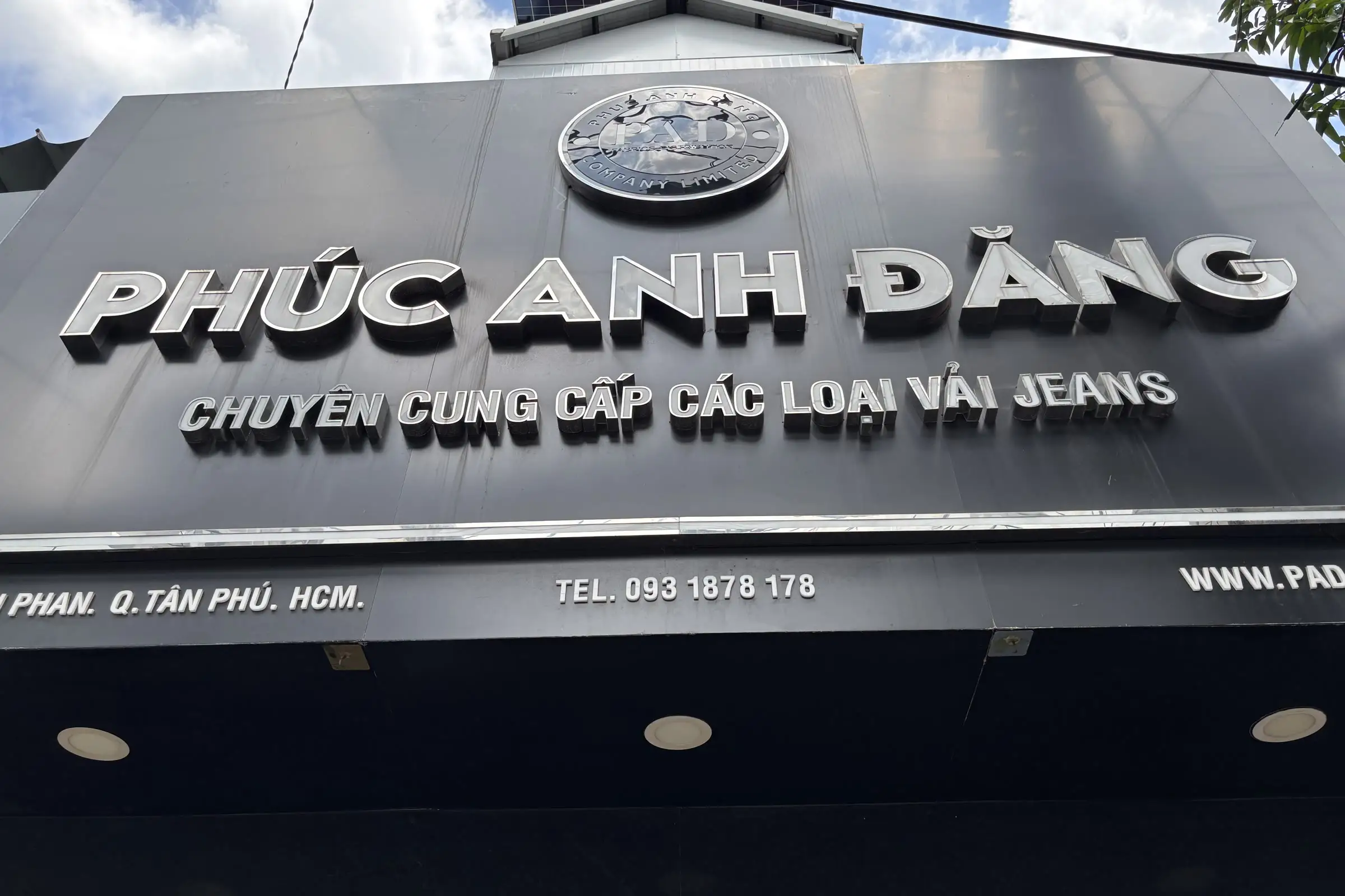 Biển hiệu cửa hàng chữ nổi PHÚC ANH ĐĂNG, bên dưới ghi Chuyên cung cấp các loại vải jeans cùng địa chỉ Quận Tân Phú và số điện thoại