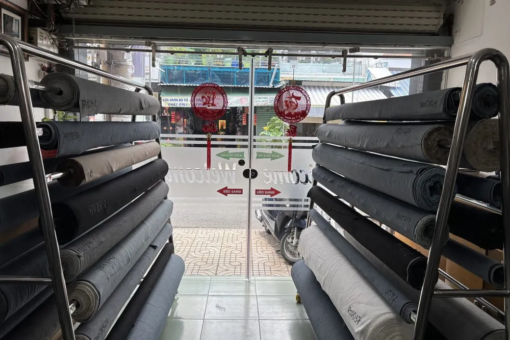Các cây vải jean cuộn tròn xếp chồng trên hai giàn kệ inox trong cửa hàng, nhìn ra cửa kính