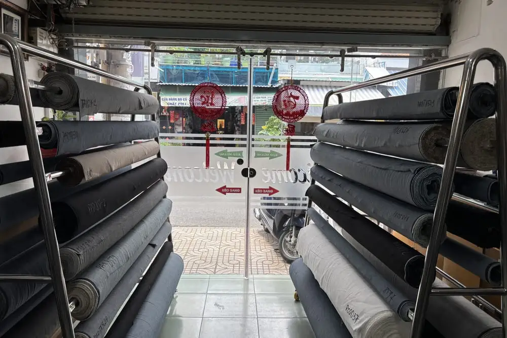 Hai giàn kệ inox xếp đầy cây vải jean xanh, xám và đen, mỗi cây ghi mã hàng bằng bút trắng, nhìn ra cửa kính cửa hàng Phúc Anh Đăng