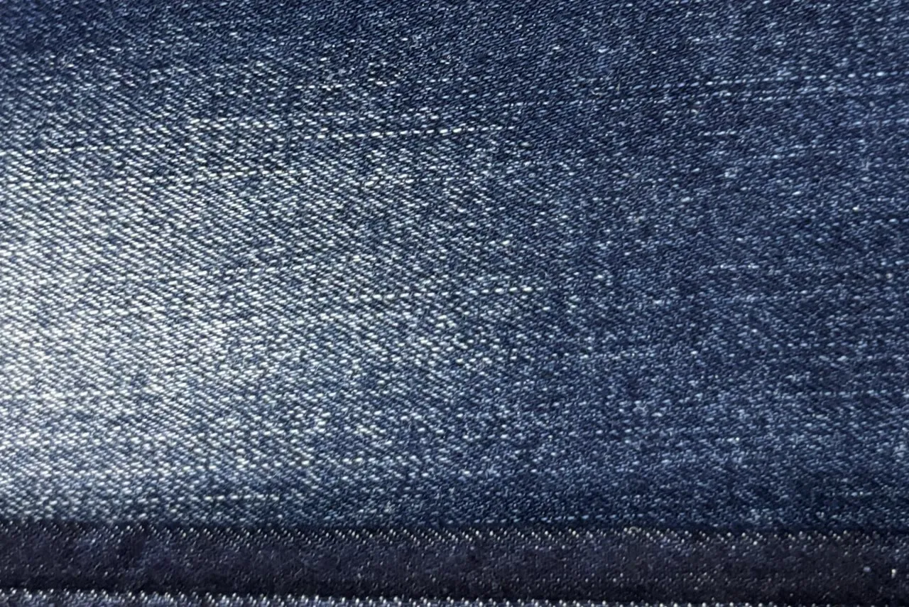 Cận cảnh mặt vải denim xanh chàm, một mảng bạc sáng lan giữa tấm, thấy rõ từng chấm sợi trắng nổi lên trên nền xanh