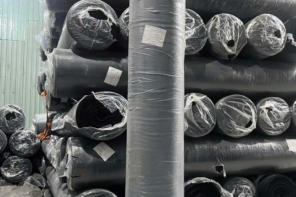 Hàng chục cây vải jean bọc nilon xếp chồng nhiều tầng trong kho, mỗi cây dán một mẩu tem giấy nhỏ ghi thông số