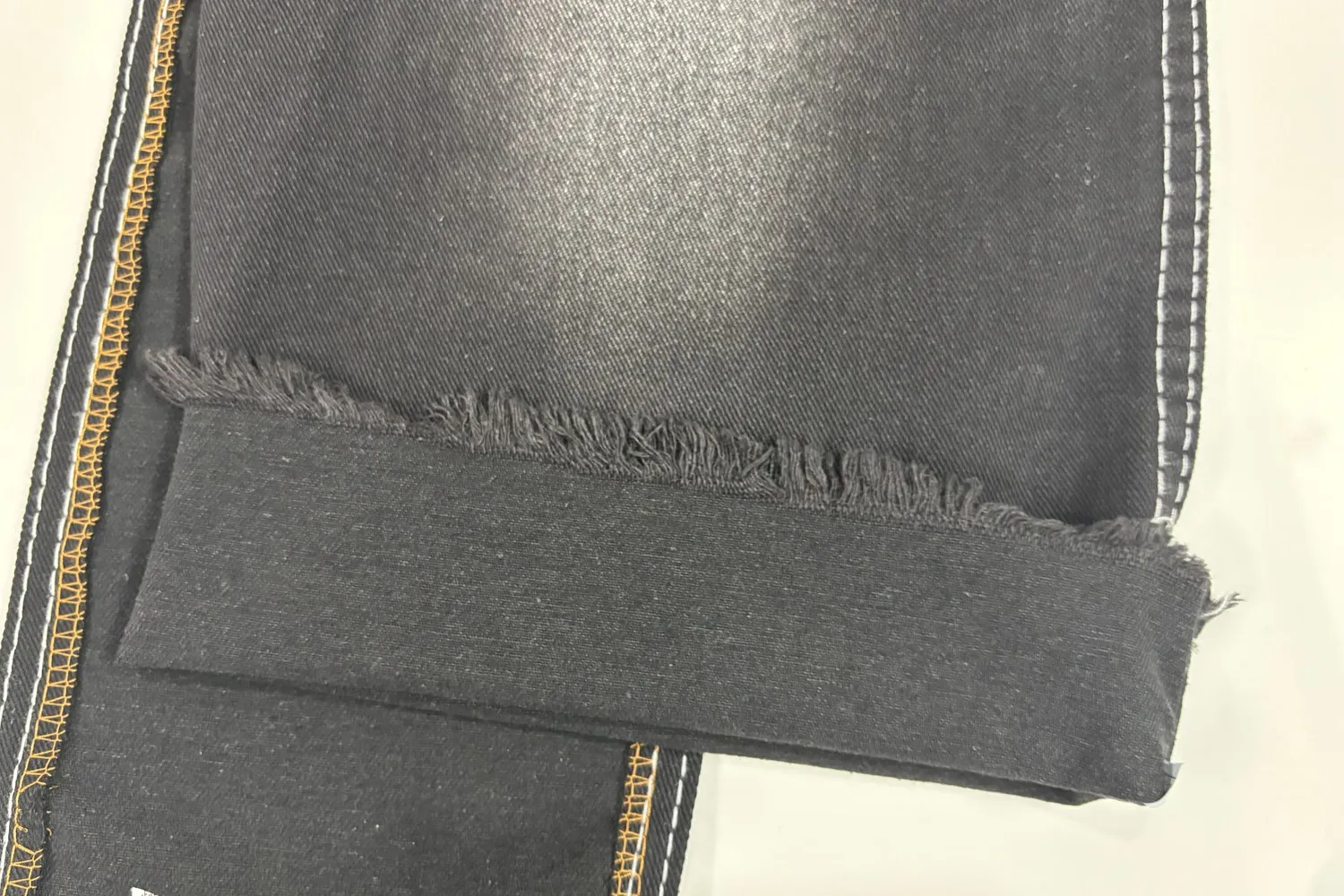 Một mảnh vải denim xám gập đôi trên nền trắng, mép cắt để tự nhiên đang tua sợi, bên cạnh là đường biên vắt sổ chỉ vàng