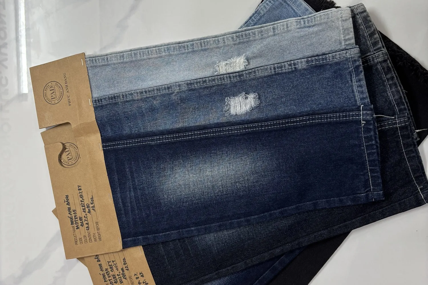 Ba mẫu vải denim xanh từ nhạt tới đậm đặt sát mép nhau trên nền trắng, mỗi mẫu kẹp một tag giấy kraft ghi thông số