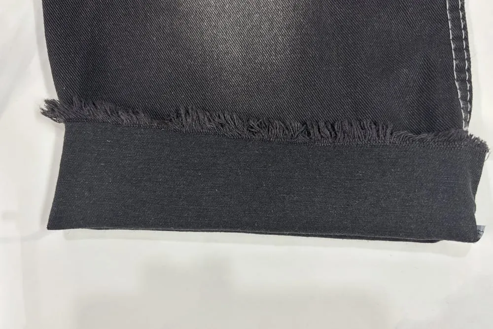 Mép cắt của một tấm vải jean đen bị tưa thành từng tua sợi ngắn, bên dưới là phần vải gấp đôi