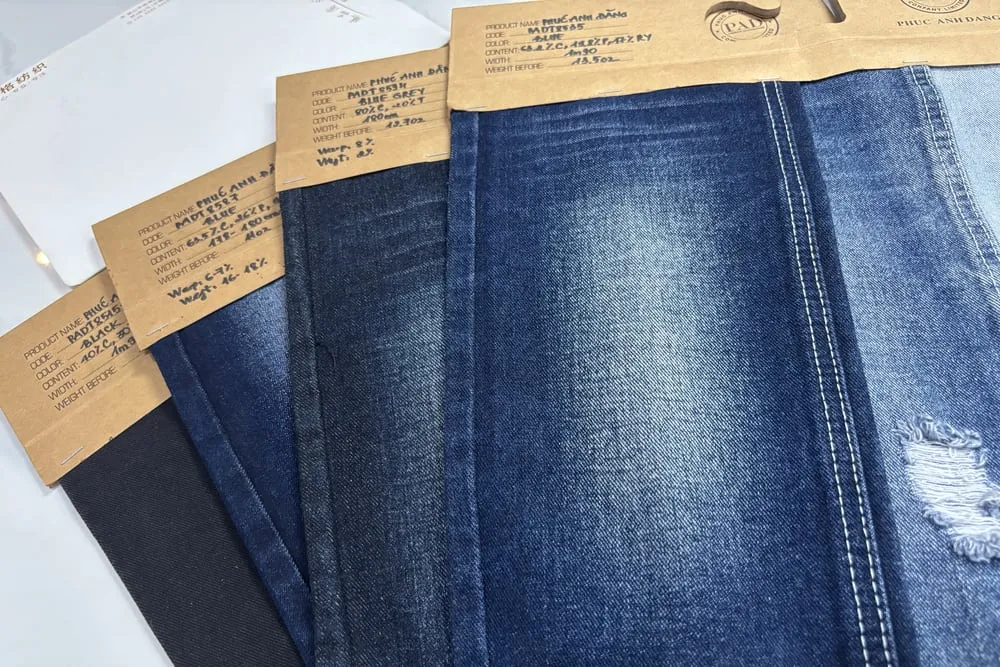 Bốn mẫu vải denim xòe chồng lên nhau, mỗi mẫu kẹp tag giấy kraft ghi tay mã hàng, thành phần, khổ và định lượng