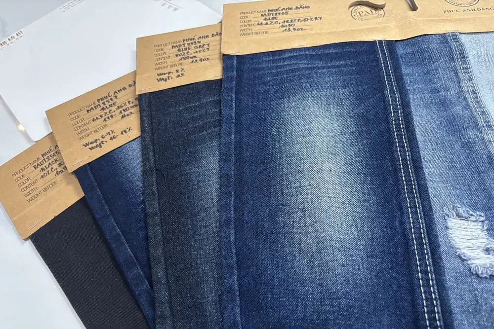 Bốn mẫu vải jean xòe ra, mỗi mẫu kẹp một tag giấy kraft ghi tay thành phần sợi, khổ vải và định lượng