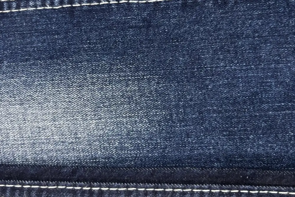 Cận cảnh mặt vải jean xanh đậm với một dải bạc sáng chạy ngang giữa tấm, hai bên là đường may chỉ trắng
